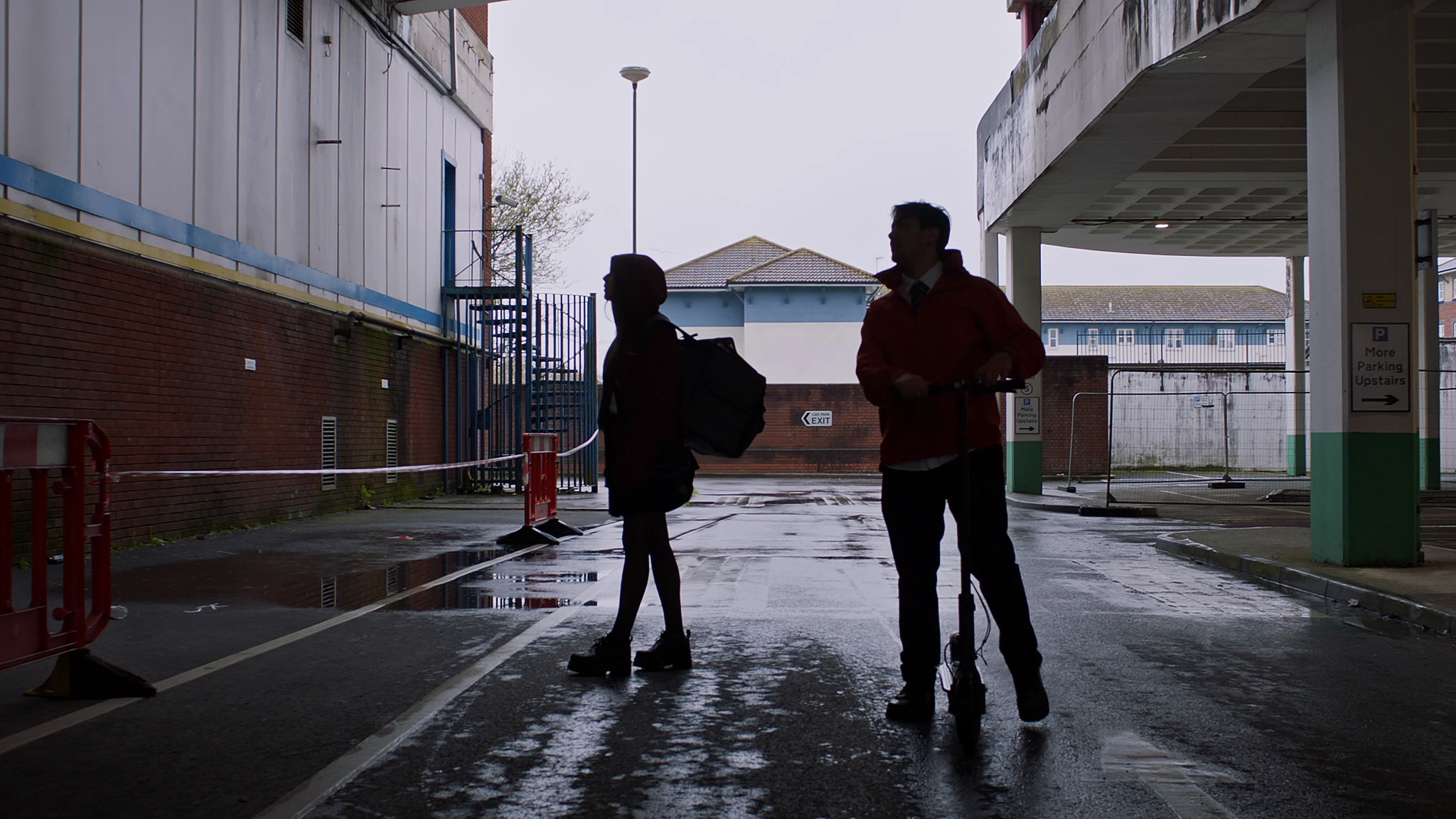 Two figures crossing a wet, empty car park
